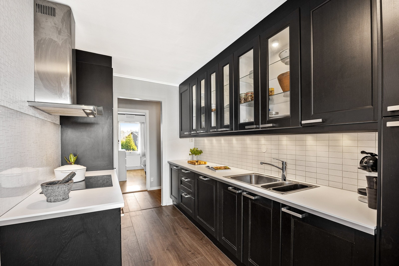 Innredningen har profilerte fronter, en laminert benkeplate og flislagt vegg mellom benkeplate og overskap. Integrerte hvitevarer som oppvaskmaskin, stekeovn, induksjonstopp, kombiskap og ventilator Galleribilde