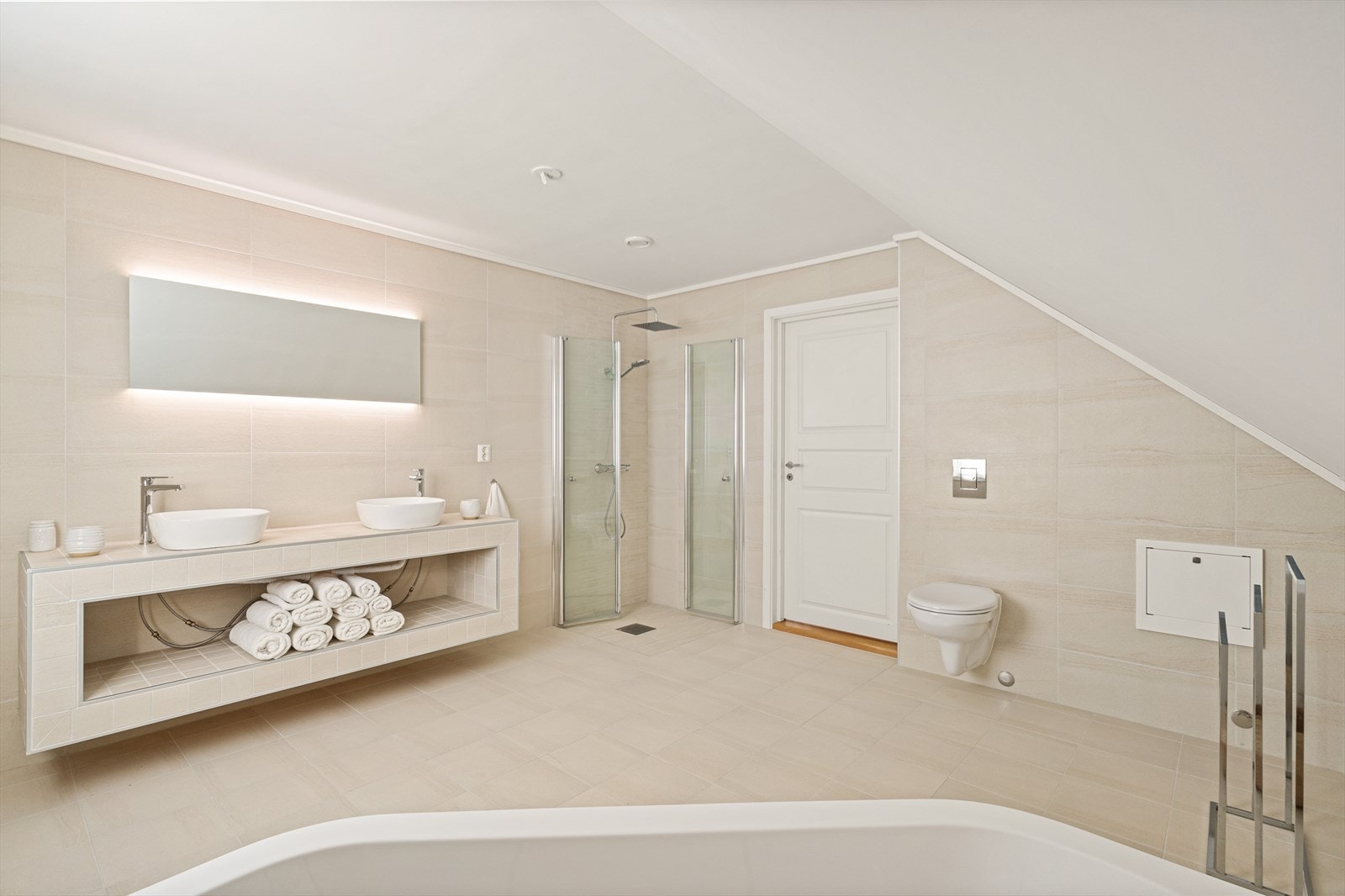 Innredningen inkluderer vegghengt toalett, dusjhjørne med glassdører, hjørnebadekar og dobbel servant med overhengende speil og belysning. Galleribilde