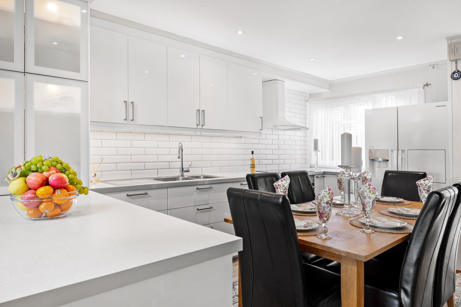 Lyst kjøkken med hvite fliser mellom benkeplate og overskap samt kjøkkenhalvøy. Galleribilde