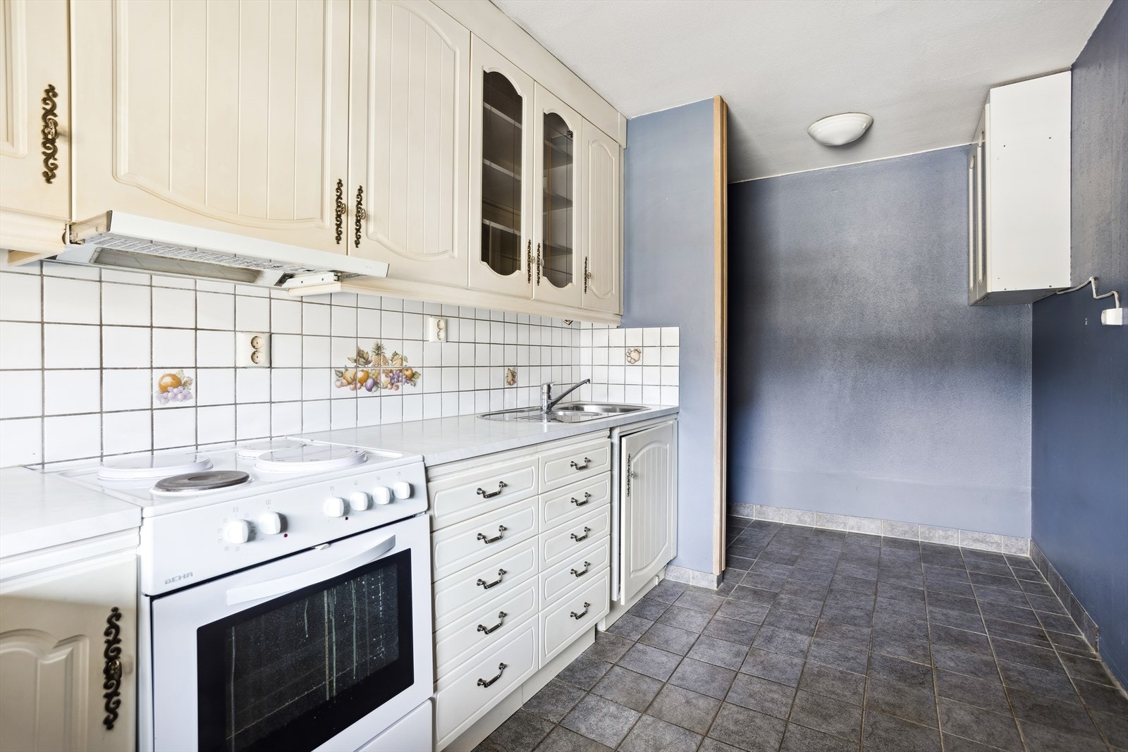 Eldre kjøkkeninnredning med profilerte fronter, to fronter av glass. Benkeplater av laminat og vask av stål. Fliser på vegg over benk. Åpning for komfyr. Galleribilde