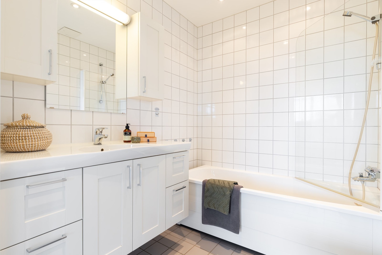 Badet har vegghengt toalett, badekar med dusj, servant med servantinnredning med dører og ovenpåliggende servant med ett-greps armatur. Galleribilde