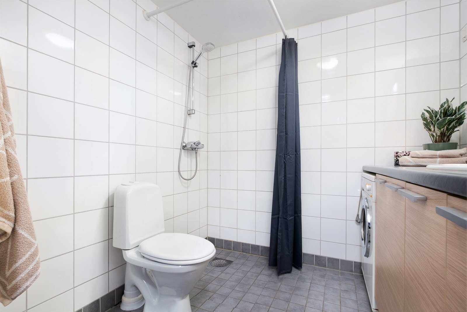 Badet er innredet med toalett, innredning med servant, opplegg for vaskemaskin (Kran og avløp), åpen dusjsone med dusjgarnityr på vegg og dusjforheng Galleribilde