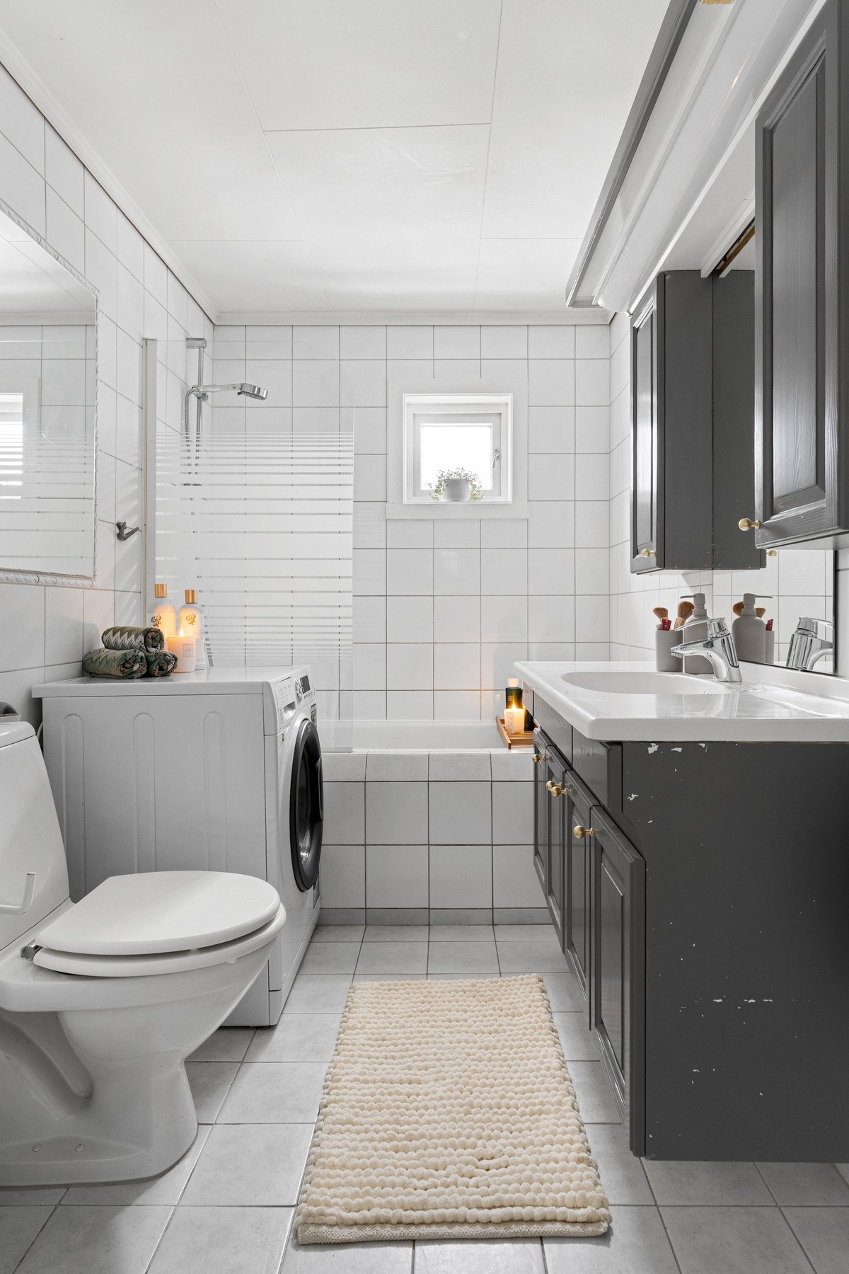 Lyst flislagt baderom utstyrt med badekar, varme i gulv, dusjgarnityr, baderomsinnredning, opplegg for vaskemaskin og toalett. Galleribilde