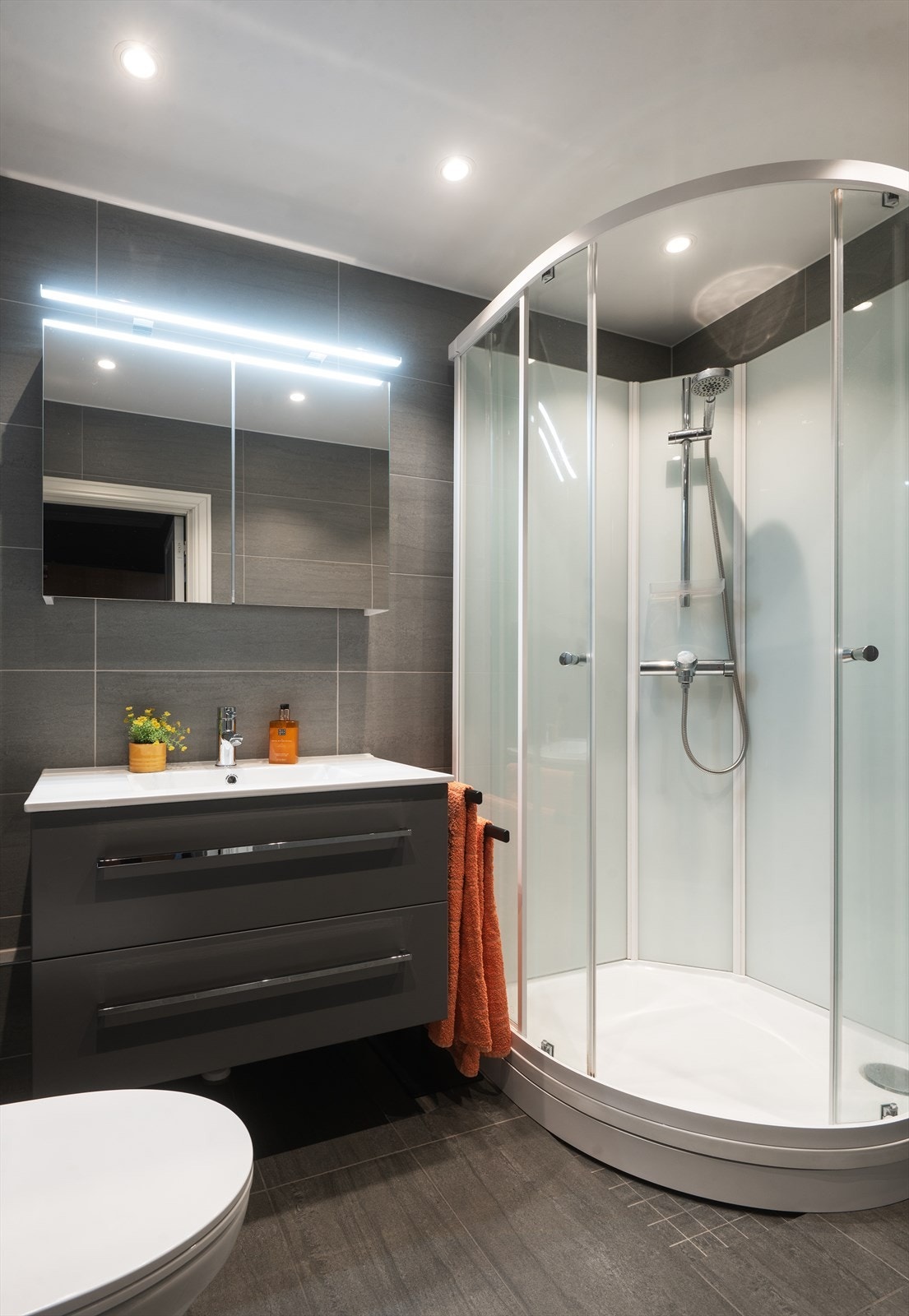 Badet er utstyrt med vegghengt toalett, servant med ettgreps blandebatteri, dusjkabinett, speilskap med belysning og vegghengt skap Galleribilde