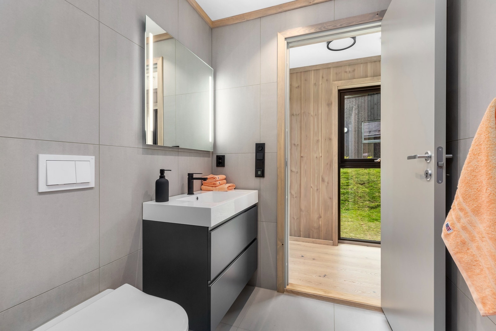 Badet er praktisk og delikat innredet med flislagt gulv og vegger. Heldekkende servant i seksjon med skuffer, speil med integrert belysning, wc med innebygget cisterne, dusjhjørne og opplegg til vaskemaskin plassert i en nisje. Galleribilde