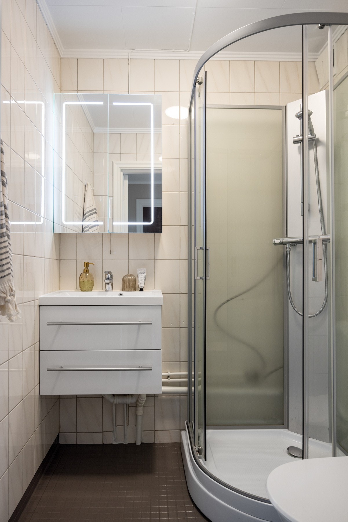 Flislagt bad med toalett, dusjkabinett i herdet glass, speilskap med integrert belysning og servantskap. Baderommet har moderniseringsbehov. Galleribilde