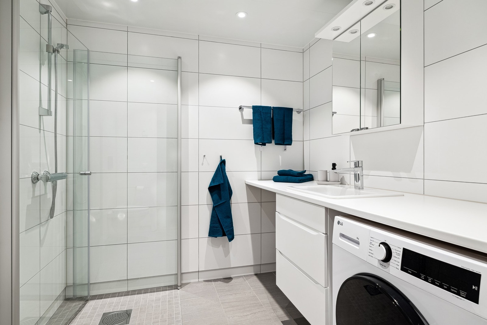 Badet har dusjhjørne med glassdører, vegghengt toalett, vegghengt servant med underskap, speil med belysning og praktisk opplegg for vaskemaskin og tørketrommel under benkeplaten. Galleribilde