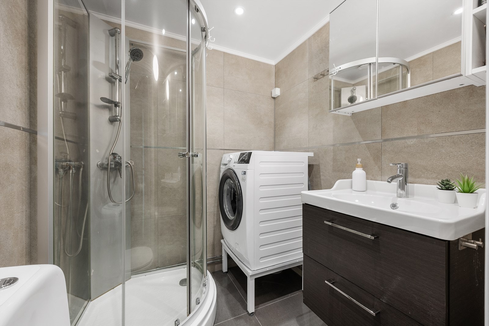 Lyst og pent flislagt baderom. Innredningen består av dusjkabinett, servant og klosett, samt benkeskap med glatte fronter. Galleribilde