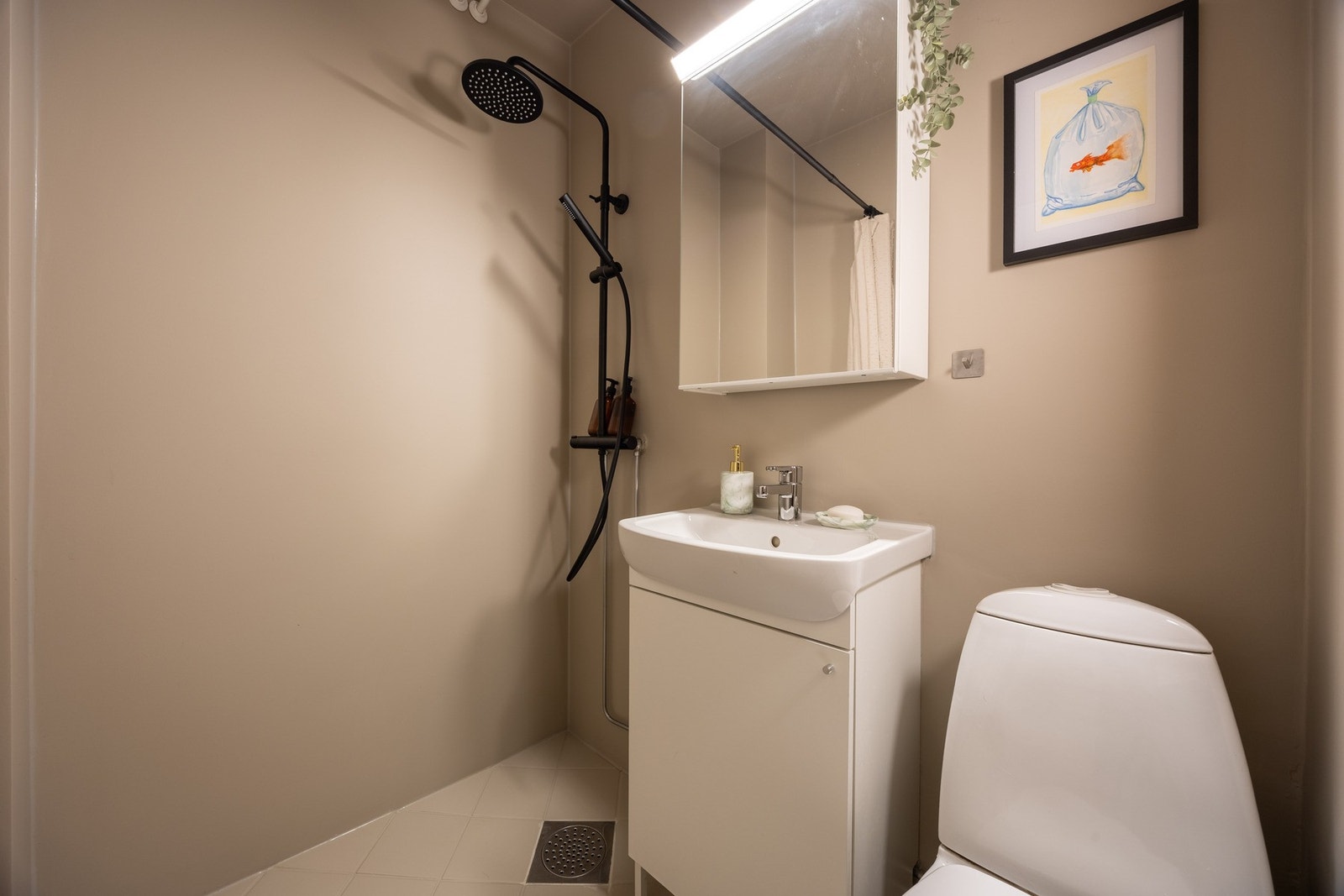 Badet har dusjhjørne, vegghengt skap med ovenpåliggende servant, speilskap, samt toalett. Galleribilde