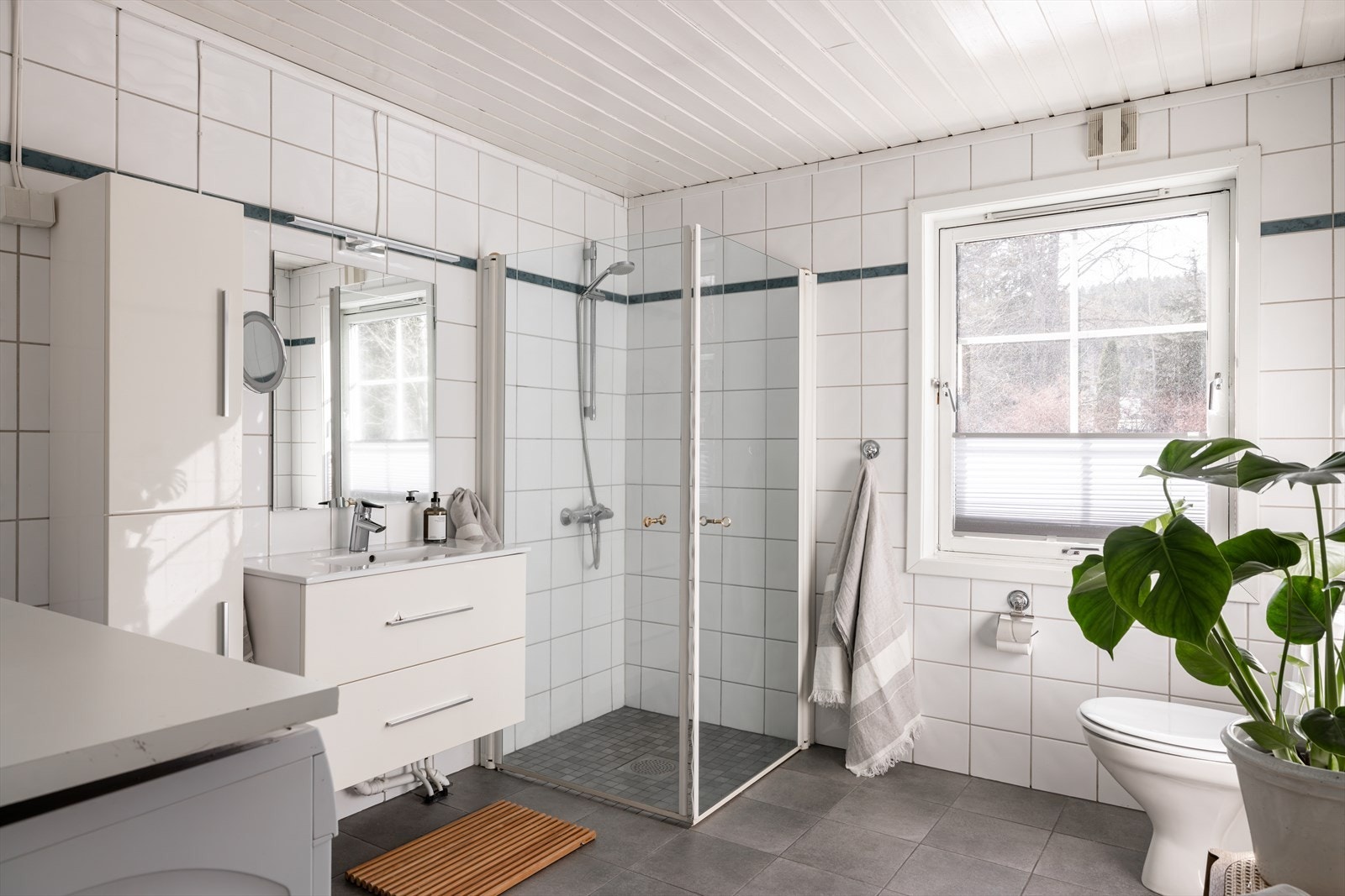 Badet har en praktisk innredning med servantskap, veggskap, speil med belysning, gulvmontert klosett og dusjsone med innfelte dusjdører i herdet glass. Galleribilde