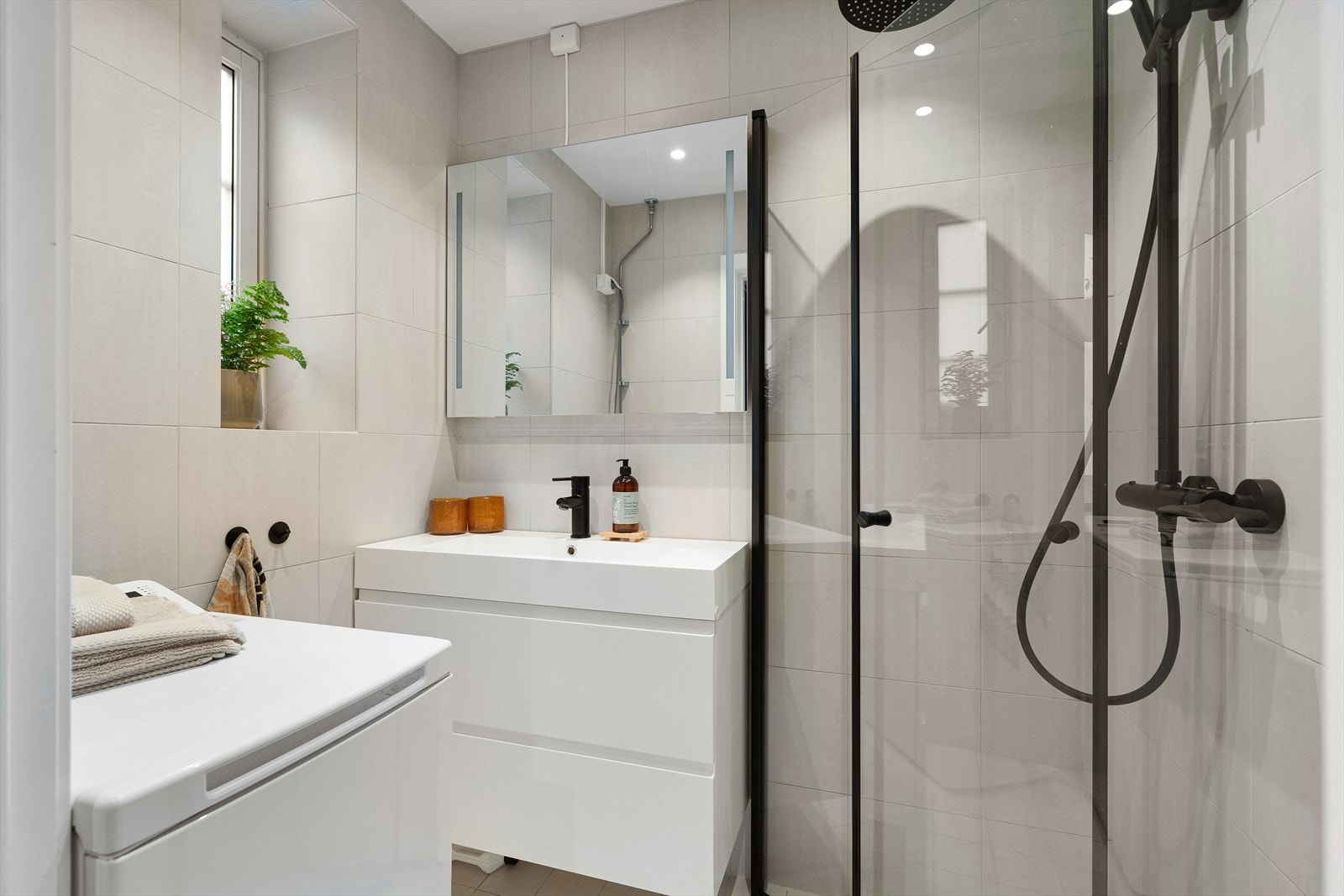 Flislagt bad med dusjplass med dusjvegger, servant med ettgreps blandebatteri og opplegg for vaskemaskin. Galleribilde