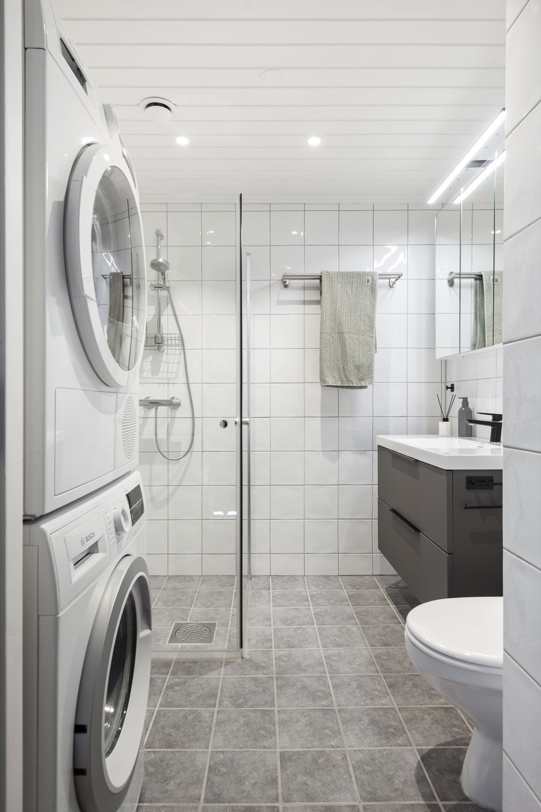 Pent baderom med fliser på gulv med gulvvarme, fliser på vegger og malt himling med downlights. Baderomsinnredningen er fra 2024 Galleribilde