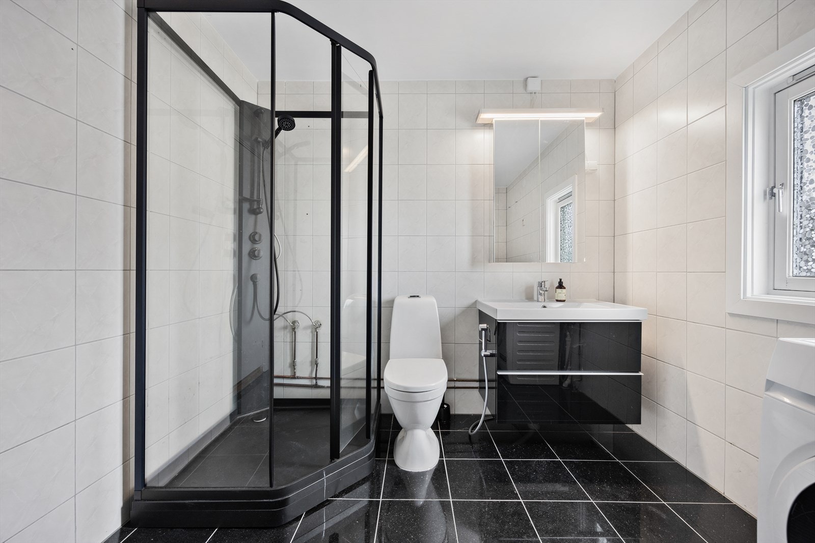 Badet inneholder servantseksjon med skuffer, overskap med speilfronter, dusjkabinett med dører i herdet glass, gulvmontert klosett, opplegg for vaskemaskin, og radiator- og relfektorovn på vegg. Galleribilde