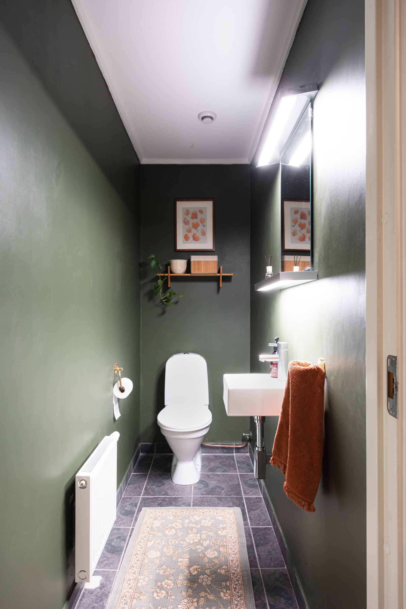 Toalettrommet har malte flater i mørke fargetoner, flislagt gulv og er innredet med servant, speil med lys på vegg over servant og et toalett. Galleribilde