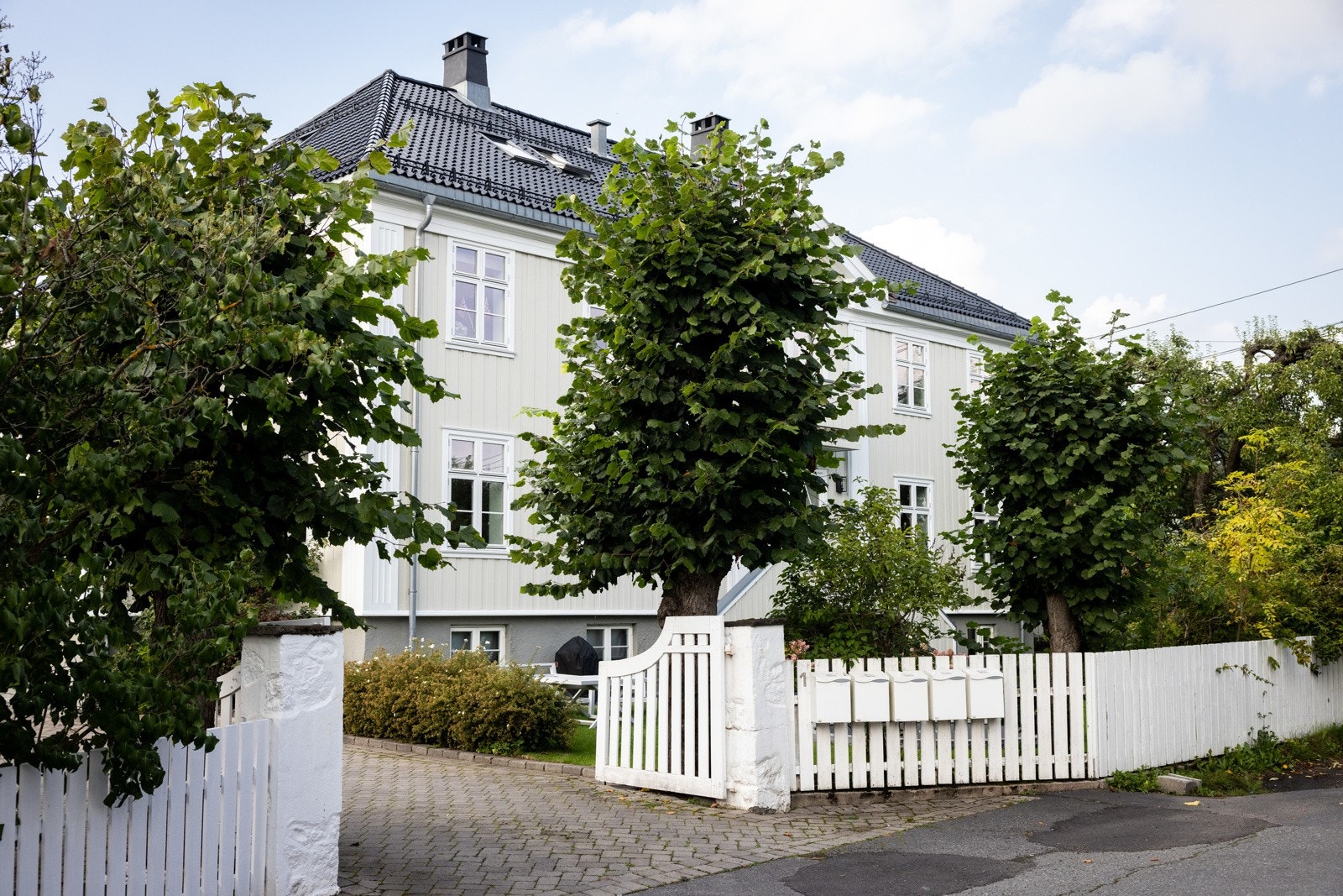Tettvokste trær, busker, hekk og et hvitmalt gjerde med tofløyet port omkranser eiendommen. Galleribilde