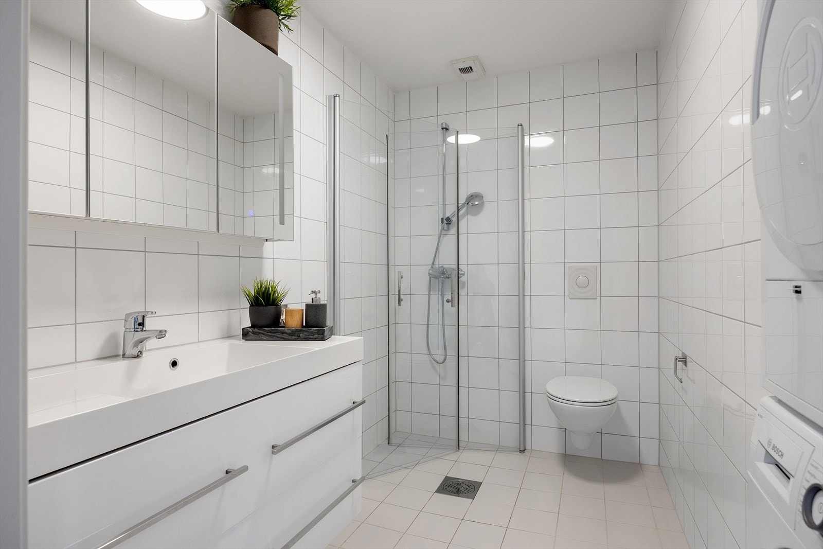 Pent flislagt baderom med gulvvarme og opplegg for vaskemaskin/tørketrommel. Badet er innredet med servantskap med servant, veggskap med speildører og innebygd berøringslys, vegghengt toalett, samt dusjsone med innfelte dusjdører. Galleribilde