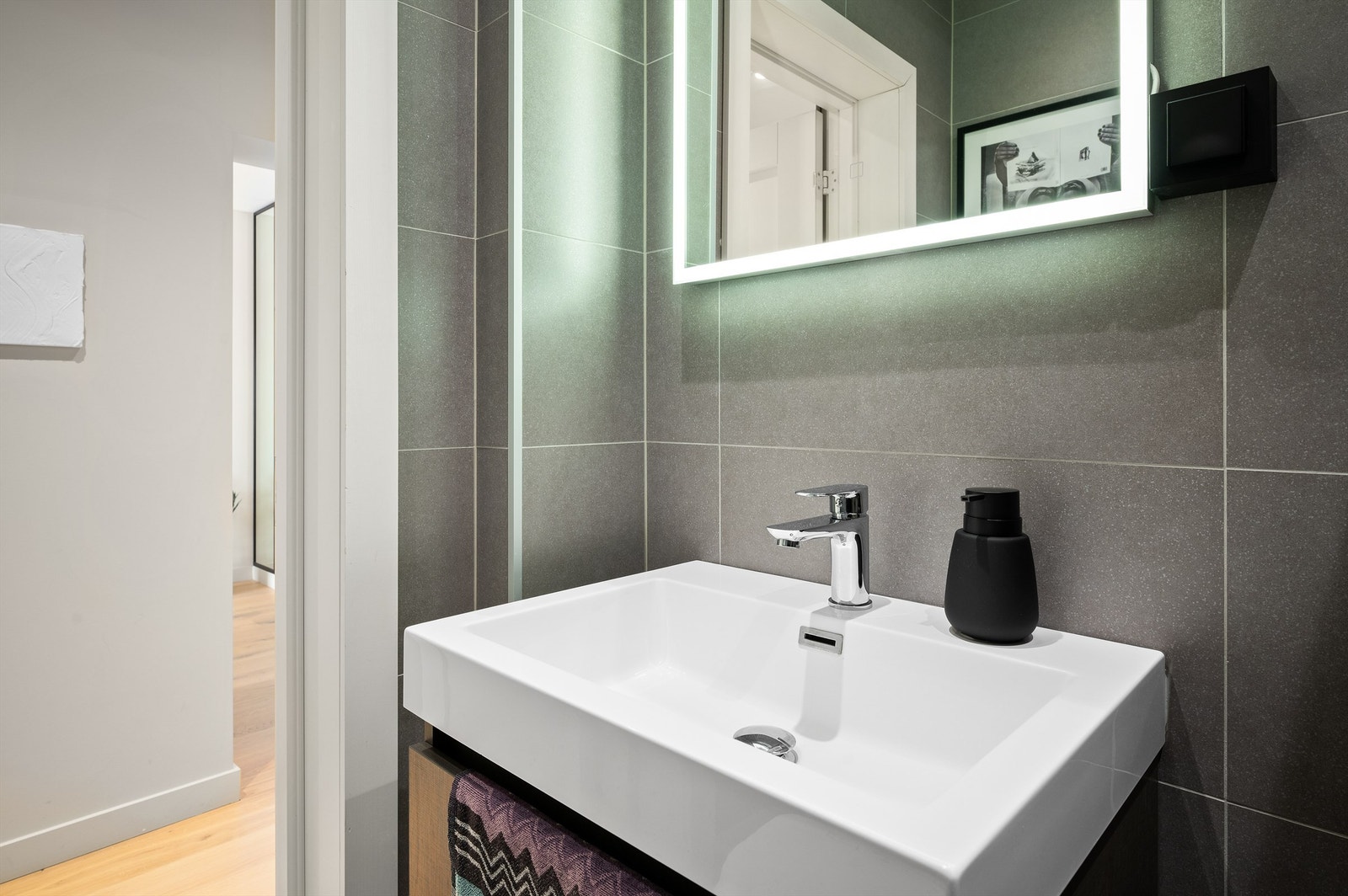 Baderomsinnredningen har grå, slette fronter og består av en heldekkende servant på en skuffeseksjon, et veggskap og et speil med integrert belysning. Galleribilde