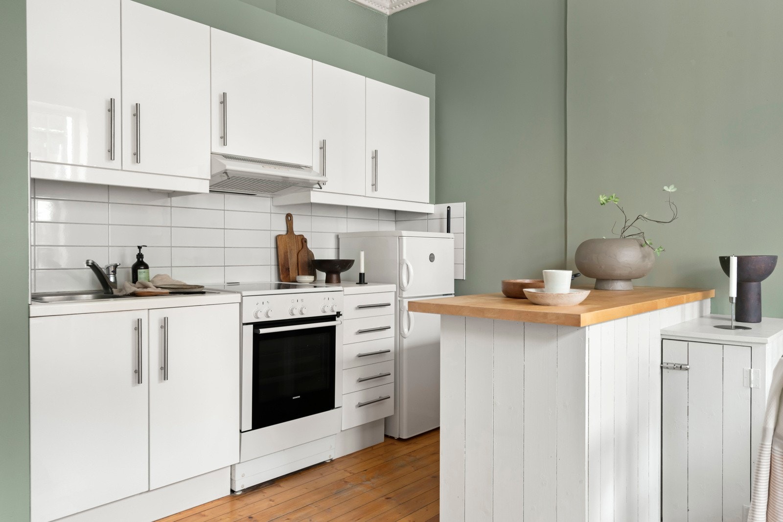 Hvit glatt, lamintert innredning med fliser mellom benkeplate og overskap, og lyslist under overskap. Det er vifte med avtrekk via mekanisk avtrekksanlegg. Galleribilde