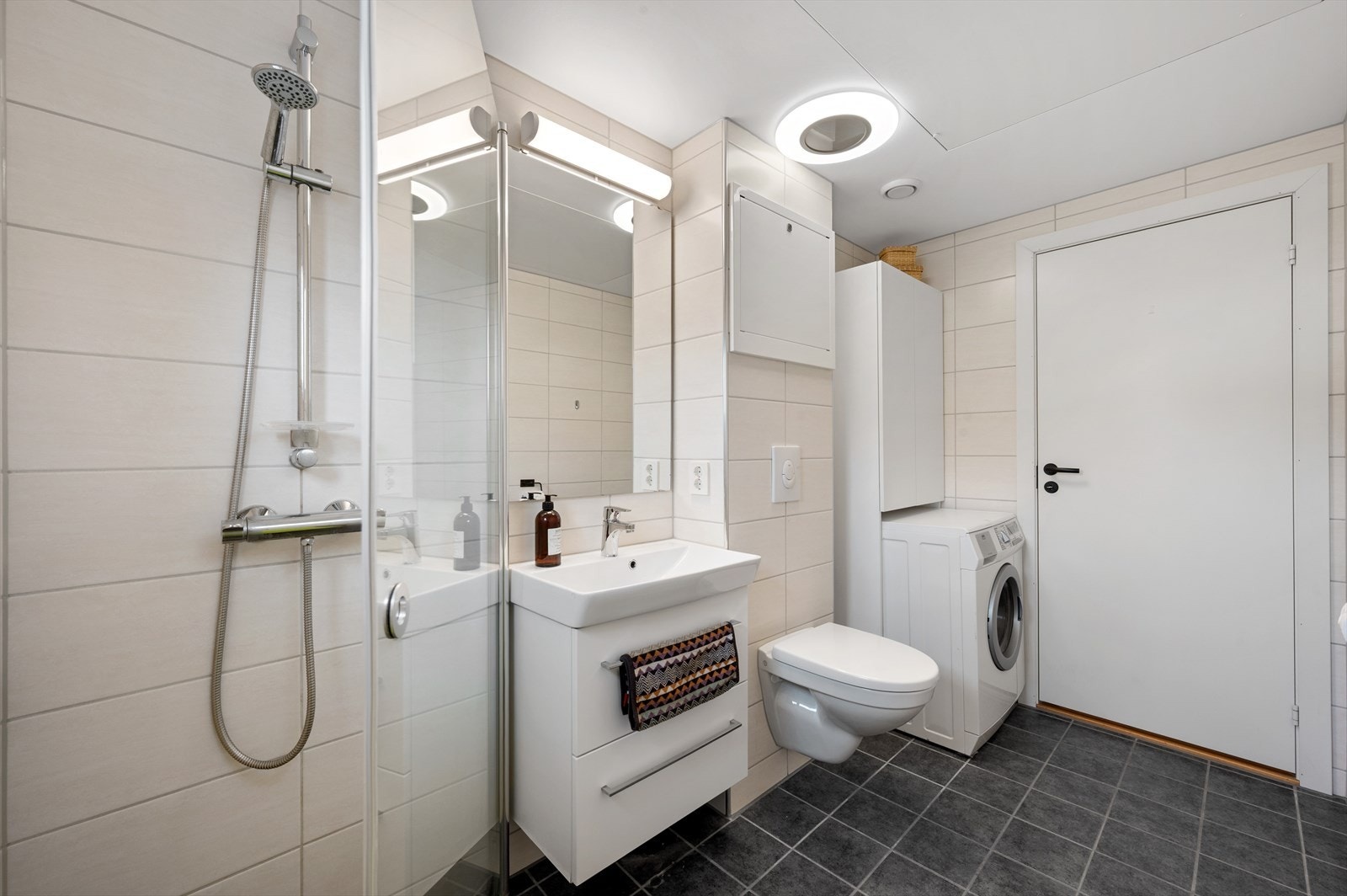 Badet er innredet med vegghengt toalett, dusjhjørne med glassdører, opplegg for vaskemaskin og servant med innredning, speil og belysning. Galleribilde