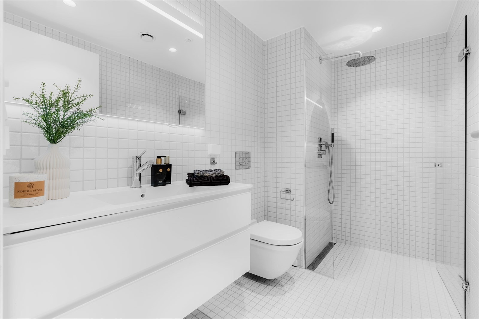 Delikat og flislagt baderom med gulvvarme. Badet er innredet med veggmonterte dusjvegger og dusj direkte på gulvet, utstyrt med termostatbatteri for enkel temperaturkontroll. Galleribilde