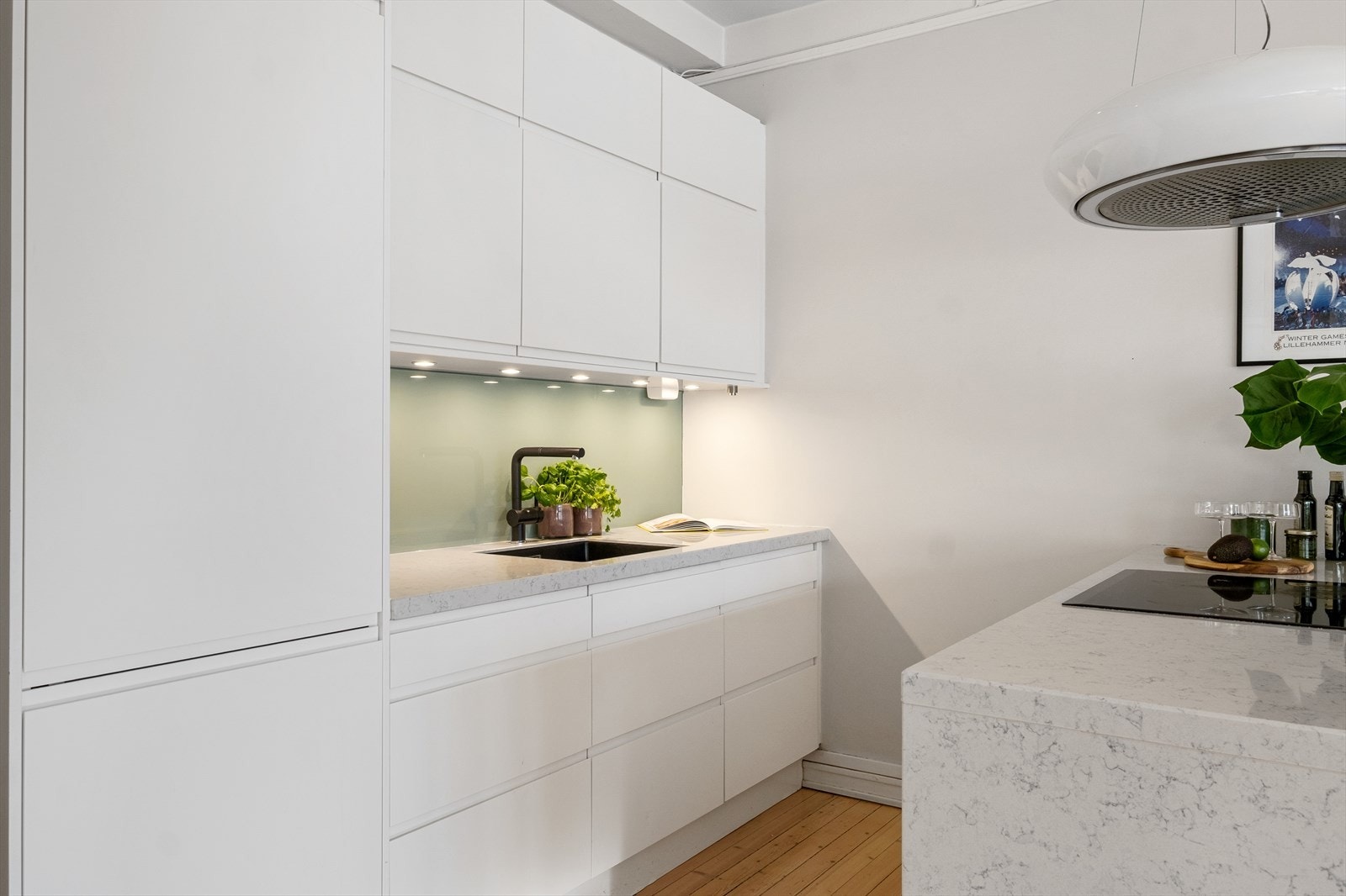 Moderne kjøkkeninnredning med glatte fronter, benkeplate av stein med underlimt vaskekum og plate og belysning over benkeplaten. Galleribilde