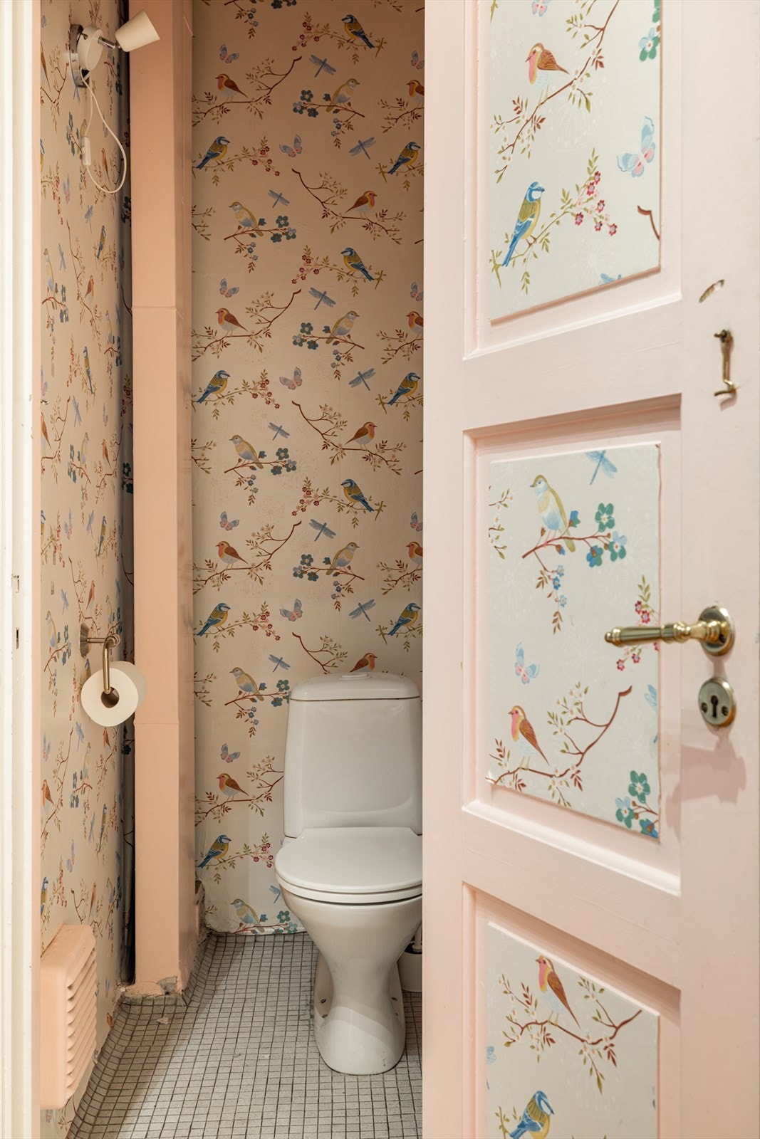 Praktisk og sjarmerende toalettrom med fliser på gulvet, mønstret tapet og malt, glatt himling. Rommet er utstyrt med gulvstående toalett og en liten servant med kaldtvannskran. Galleribilde