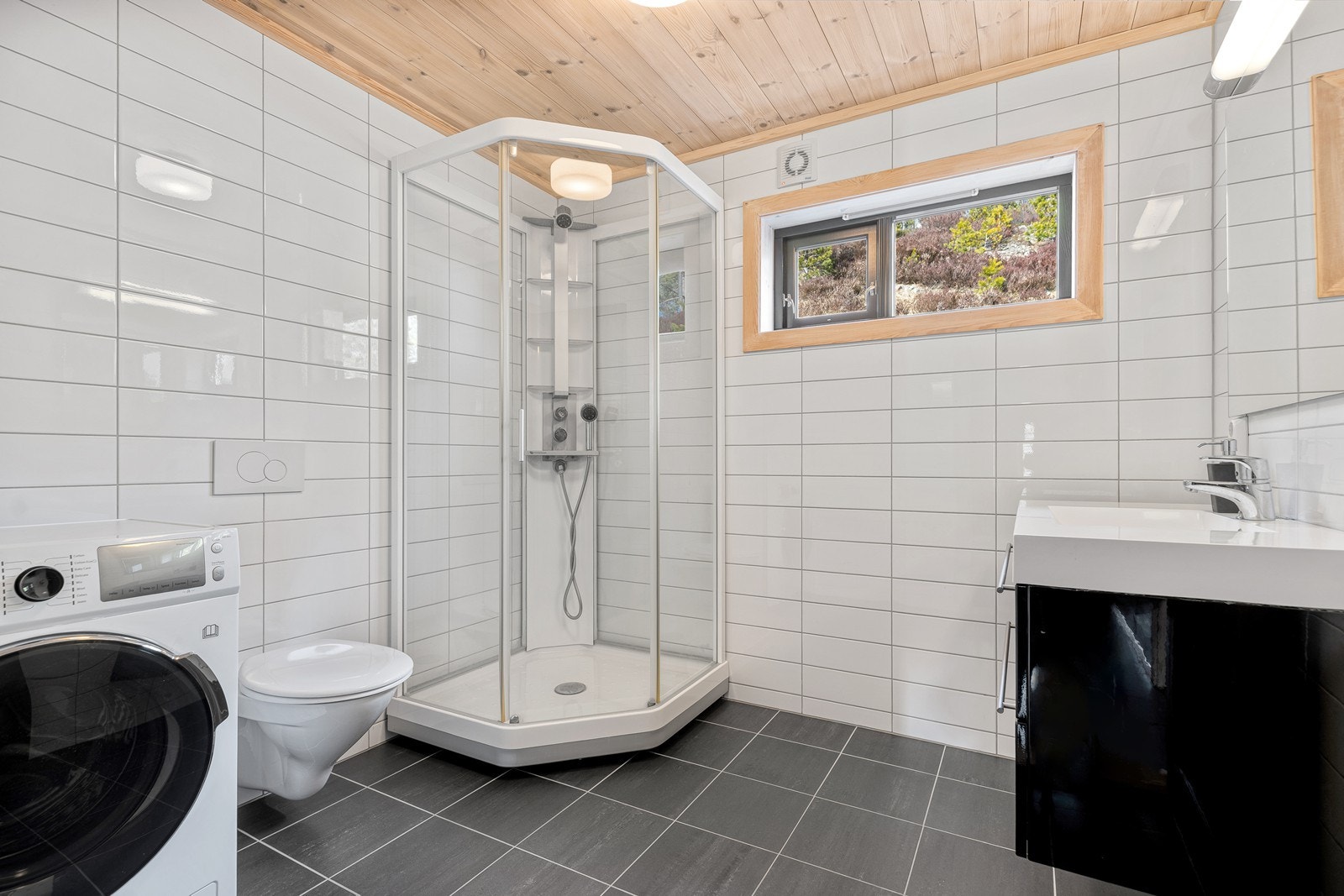 Badet er innredet med vaskemaskin, vegghengt toalett, dusjkabinett i hjørne og flott servant med speil Galleribilde