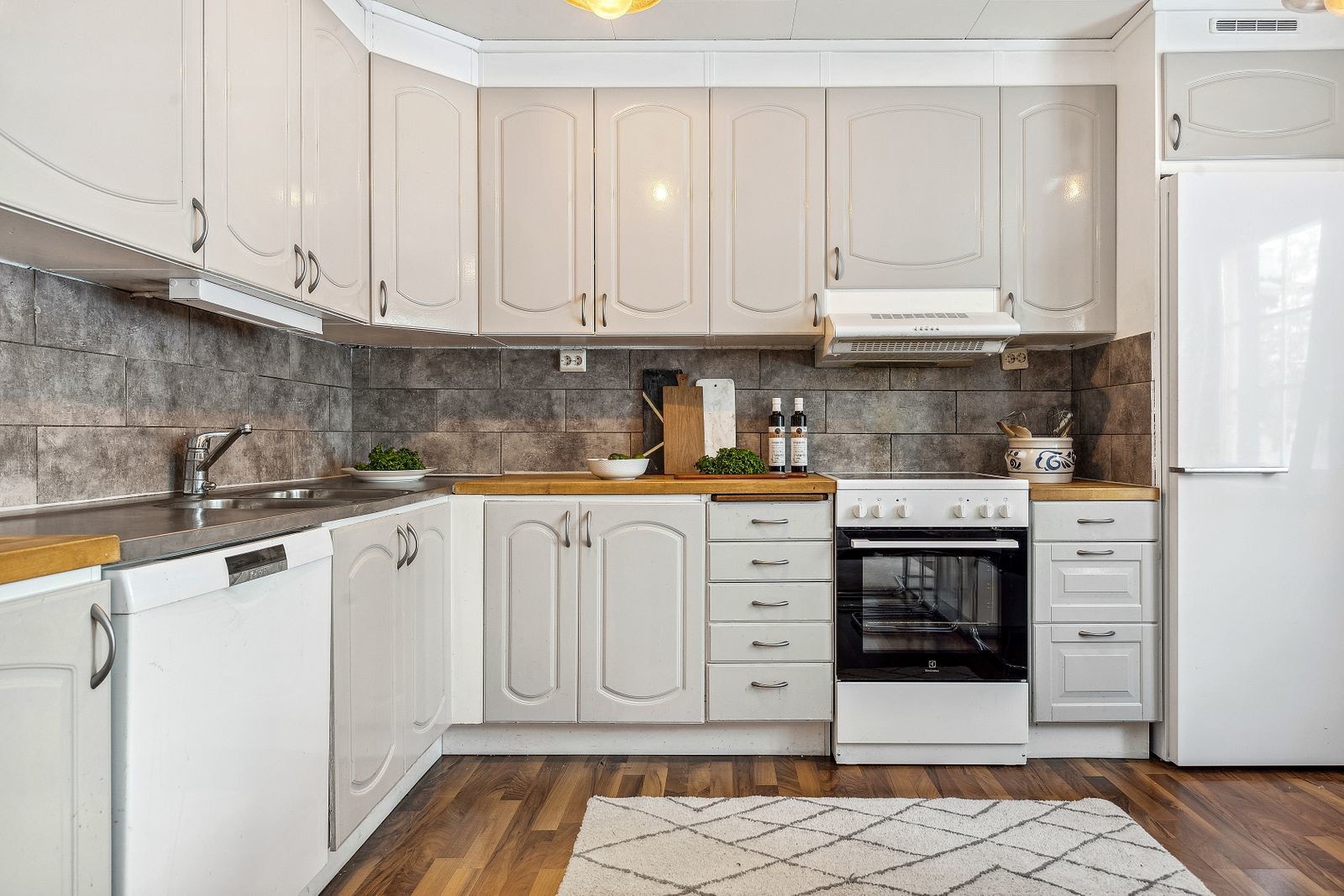 Kjøkkeninnredning med profilerte fronter. Laminat på gulv, malt trekledning på vegger og fliser over kjøkkenbenker. Benkeplater i heltre. Kjøkkenvifte med avtrekk til kanal, integrert i overskap. Galleribilde