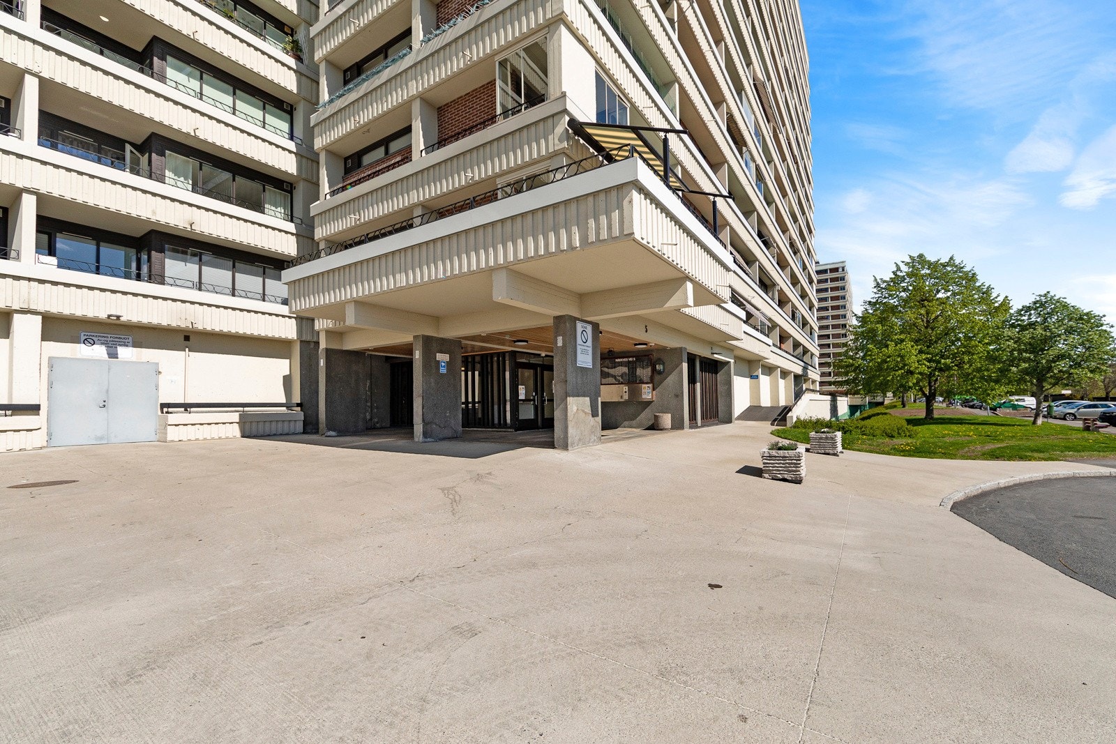 På fremsiden av blokkene er det gjesteparkering, asfalterte gangveier og flere benker omkranset av grønne busker Galleribilde