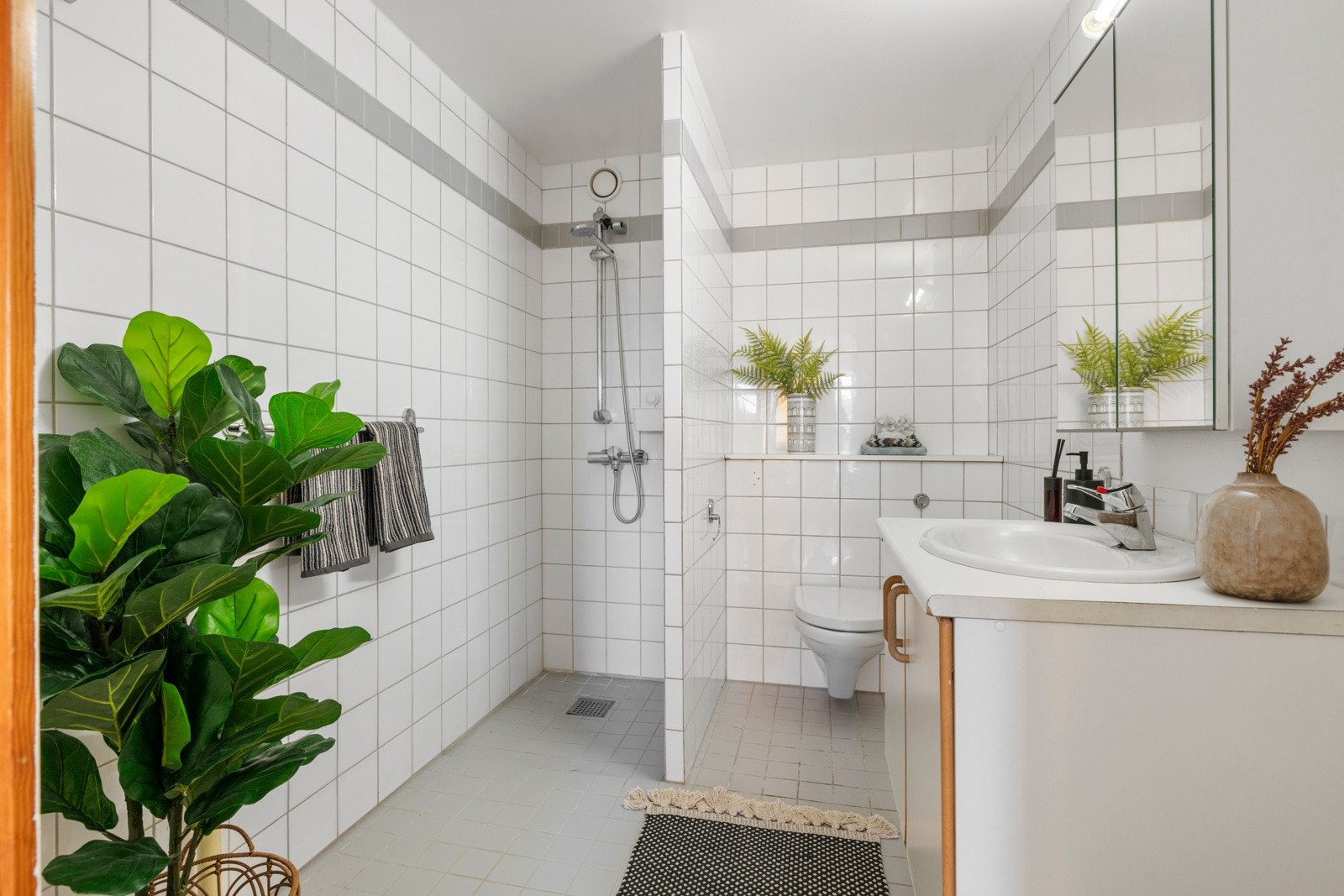 Lyst flislagt baderom med vegghengt WC, servantskap, speilskap og dusj i hjørnet. Galleribilde