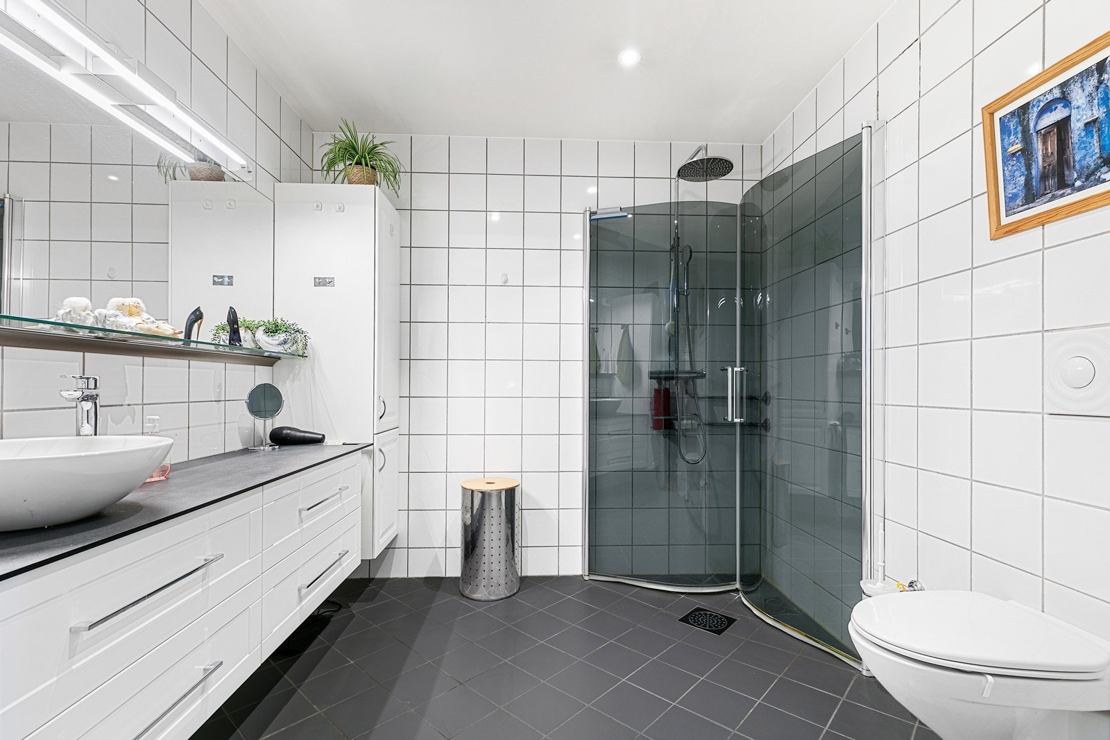 Lyst og pent, flislagt bad med varmekabler i gulv og downlights i tak. Badet er utstyrt med vegghengt toalett og dusjhjørne med innfellbare dusjvegger. Galleribilde