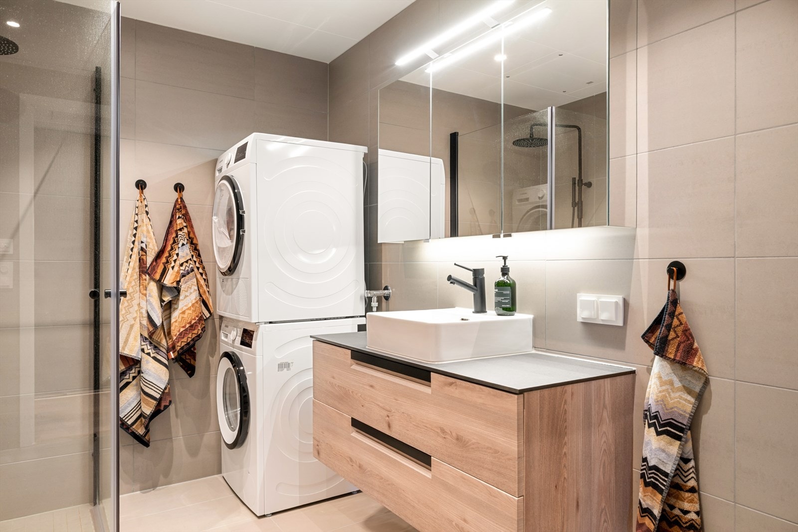 Gulvvarme og innredning som inkluderer dusjhjørne, servant med underskap, speil med belysning, vegghengt toalett og opplegg for vaskemaskin. Galleribilde