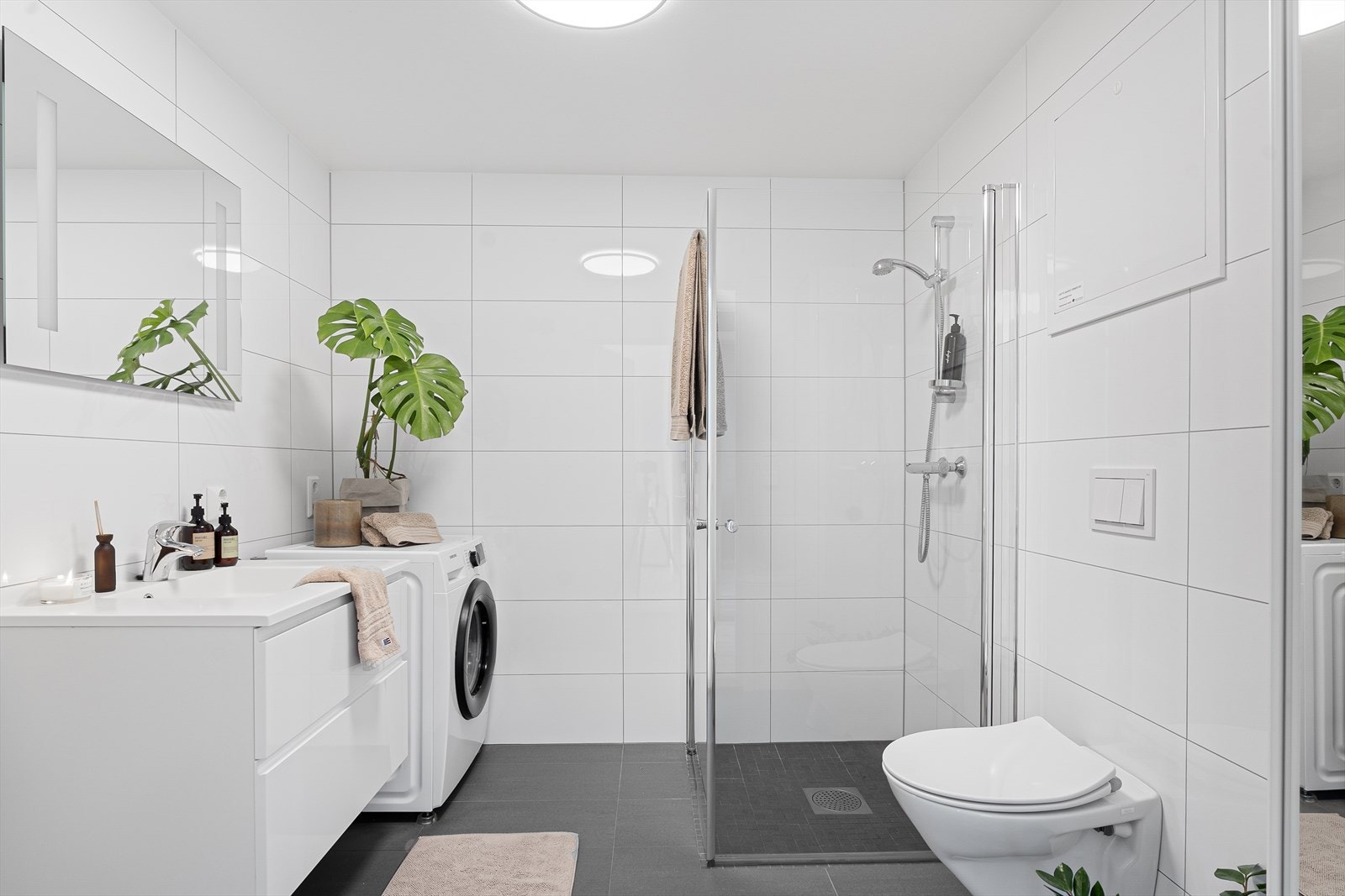 Pent, fliselagt baderom som inneholder dusjnisje, baderomsinnredning med vask og speil, vegghengt toalett og opplegg for vaskemaskin. Galleribilde