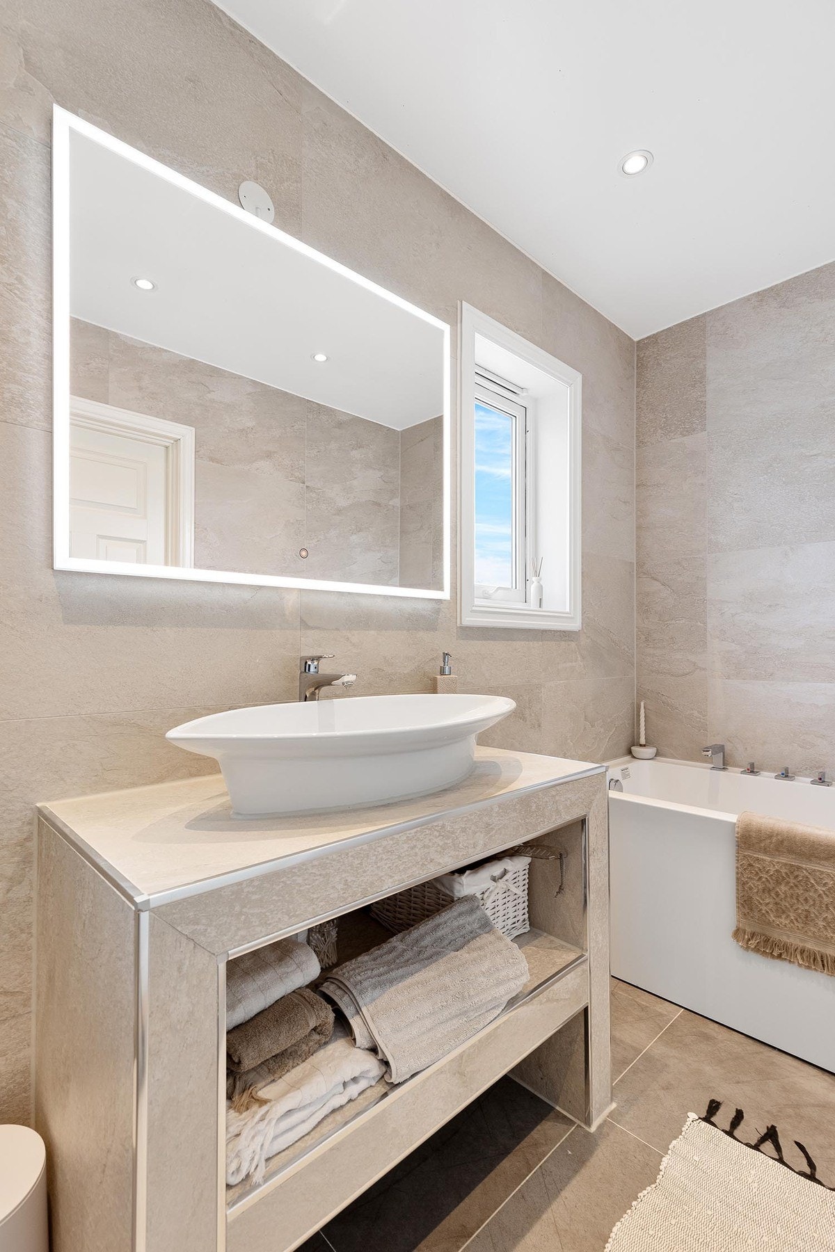 Dette delikate badet fra 2017 har lyse fliser, plassbygget baderomsmøbel med bolleservant, og et speil med integrert lys. Galleribilde