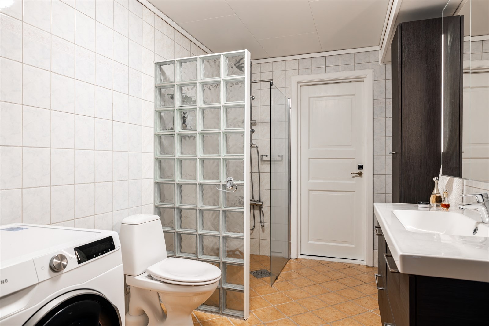 Badet er romslig og praktisk innredet, med lyse fliser på veggene og varme, terrakottafargede fliser på gulvet. Galleribilde