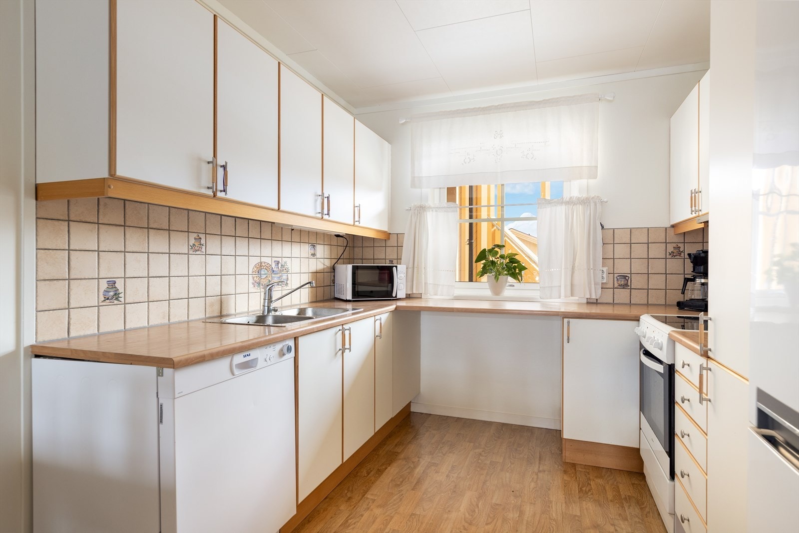 Kjøkkendelen har en hvit innredning med slette fronter, laminat benkeplate og flis over kjøkkenbenk. Galleribilde