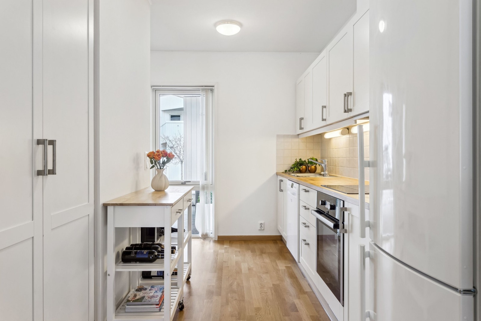Hvit kjøkkeninnredning med profilerte fronter, heltre benkeplate, kjøkkenfliser, ventilator og nisje for kjøl/frys. Galleribilde