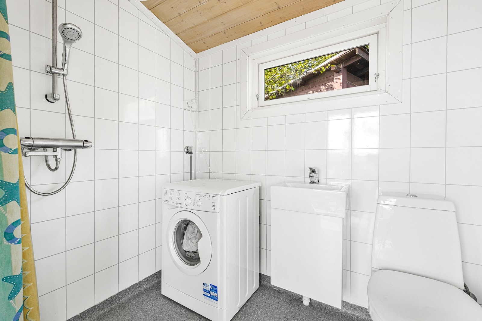 Lyst bad med vegghengt servantinnredning, dusjhjørne med forheng, gulvstående toalett og opplegg for vaskemaskin. Galleribilde