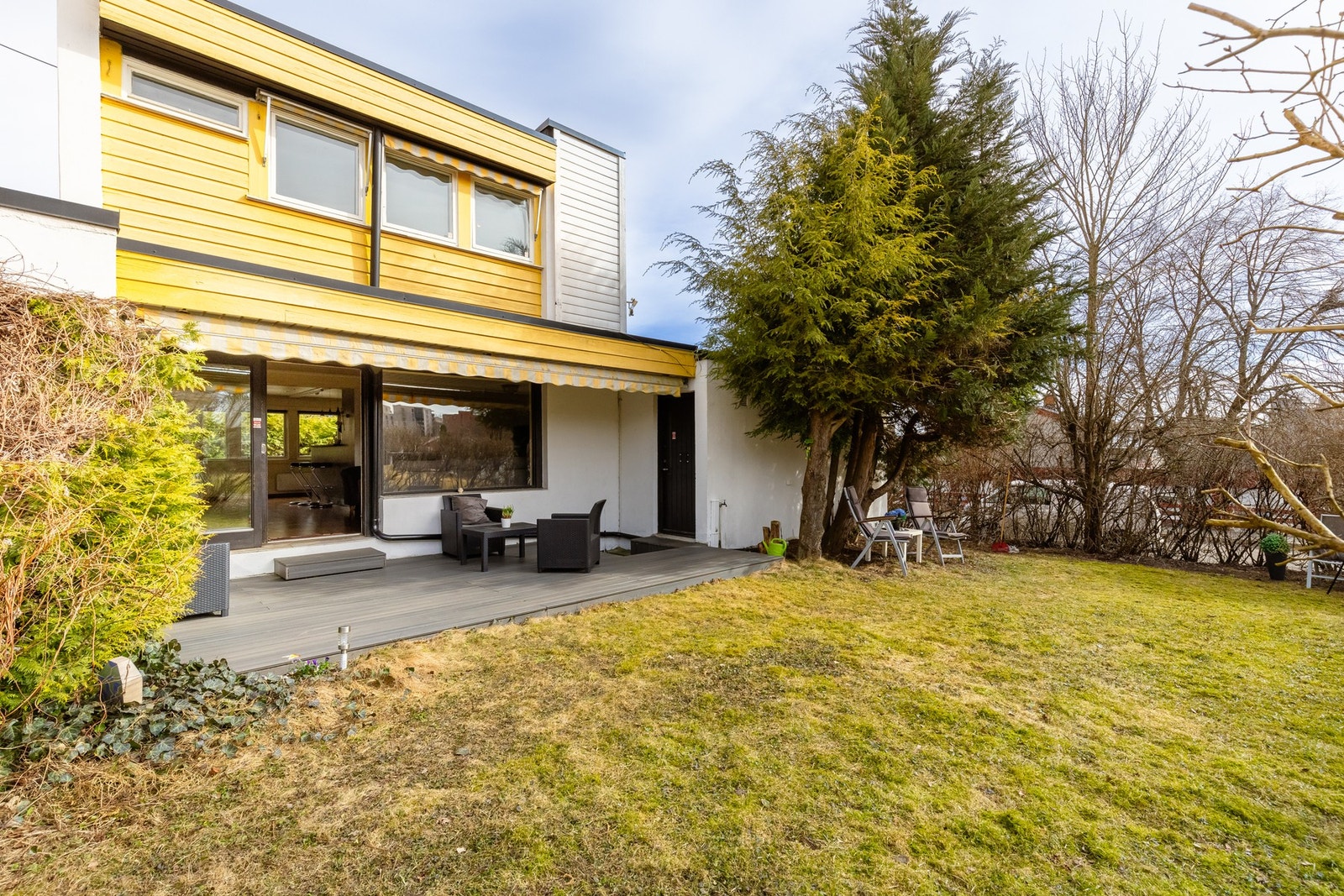 Funkishuset (1968) ligger på en hjørnetomt og vender sydvest med solrik og vedlikeholdsfri kompositt-terrasse (20 kvm, 2019). På bildet ser man dør inn til dobbeltgarasje (35 kvm) med plass til to biler og mye lagringsplass. Lettstelt hage med trær og syr Galleribilde