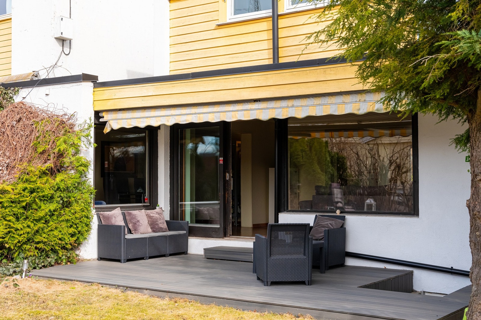 Plass til flere sittegrupper på den vestvendte terrassen. Skyvedør inn til stue. Spisestue kan ses i vindu til venstre. På høyre av bildet ses nedgang til rømningsgodkjent vindu (2018) i kjellerstue. Inngang til dobbeltgarasje til høyre. Galleribilde