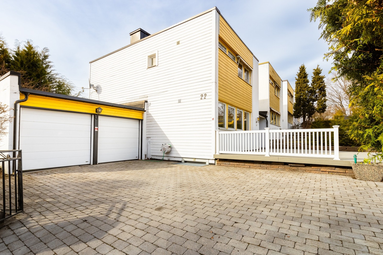 Foran huset er gårdsplassen pent opparbeidet med belegningsstein. Huset har dobbelgarasje med elbil lader og god plass til to biler. Smijernsport kan avlukke gårdsrommet. På bildet ses rist over vindu til kontor i kjeller. Galleribilde
