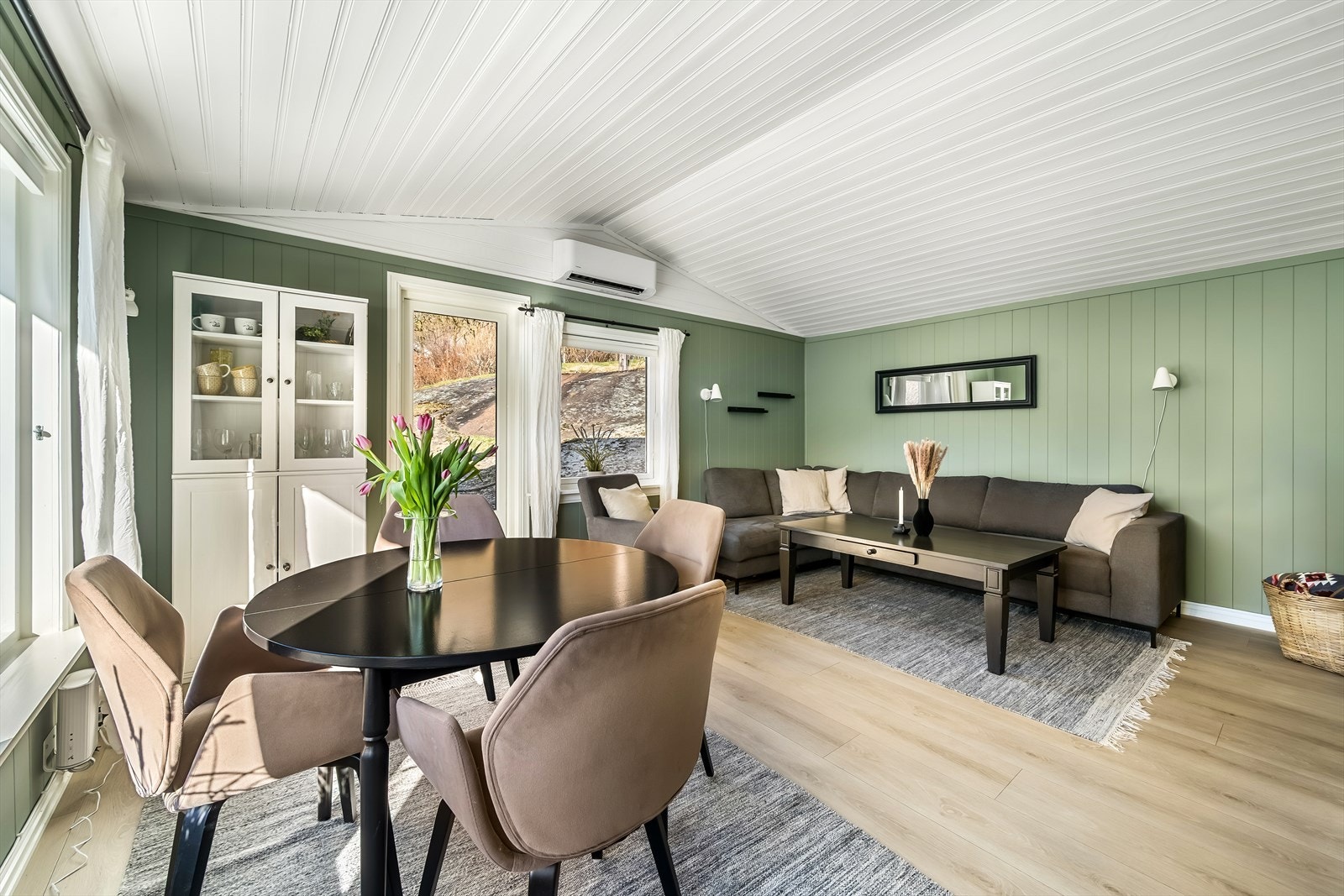 Lys og innbydende stue med store vinduer og moderne fargetoner. Varmepumpe på vegg. Galleribilde