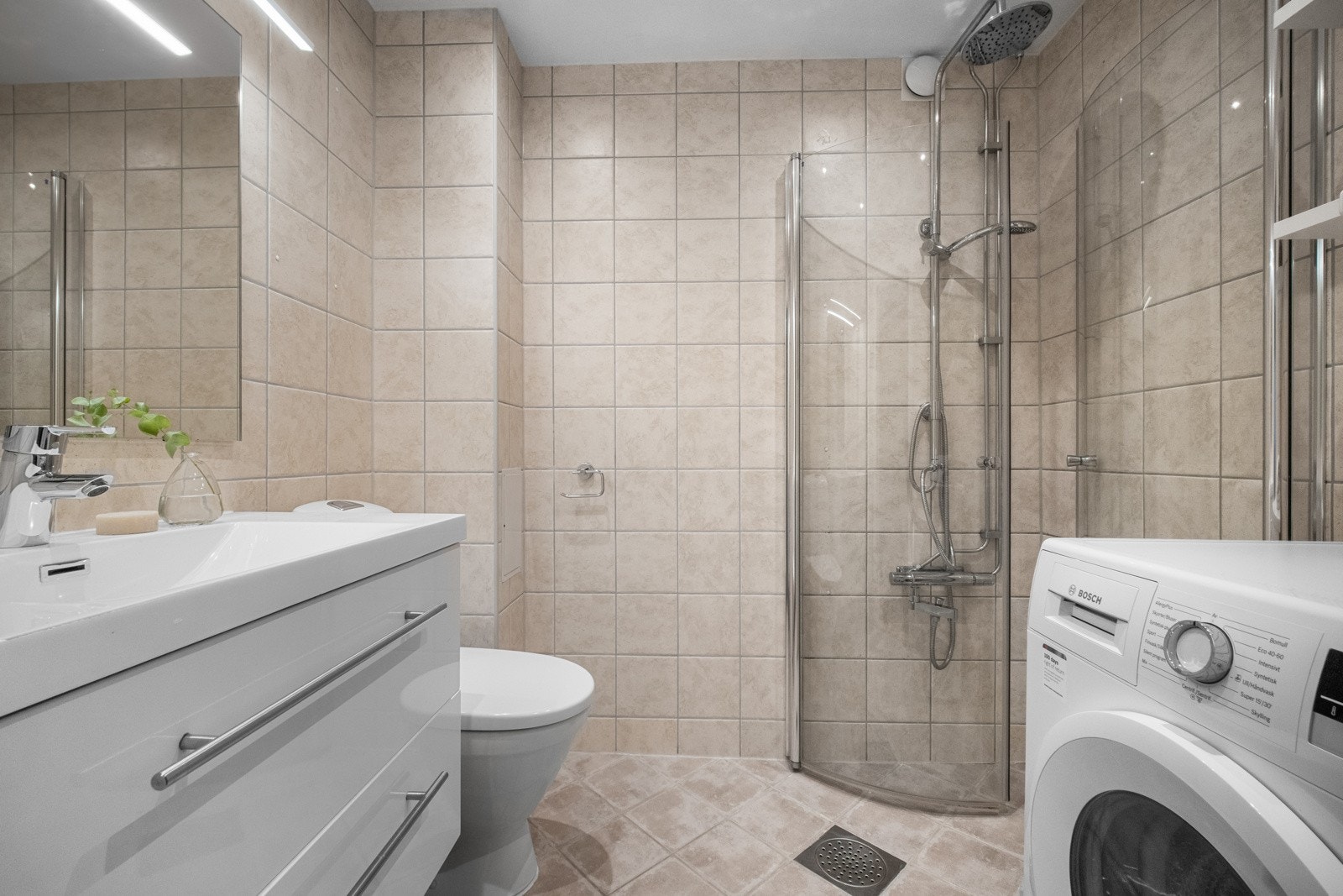 Badet er utstyrt med heldekkende servant, speil med lysarmatur, gulvmontert klosett og dusjhjørne med svingdører. Galleribilde