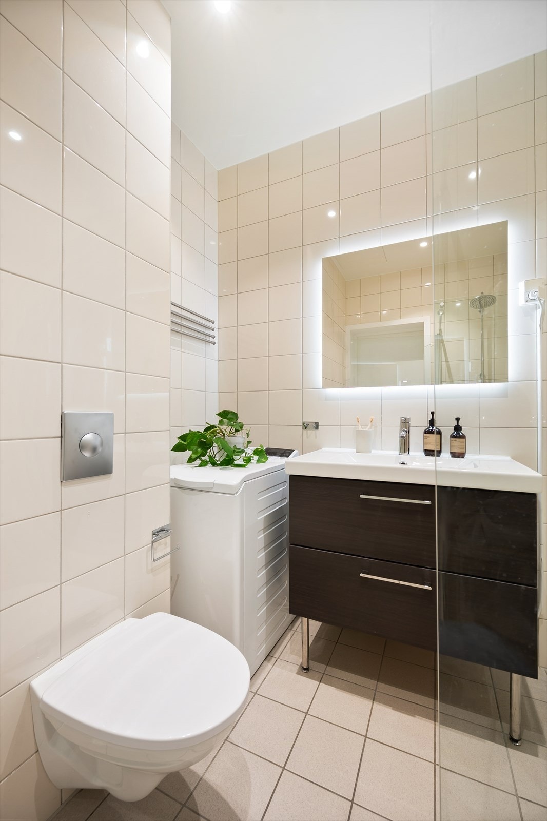 Badet inneholder veggmontert toalett, dusjhjørne med svingbare glassdører, servantskap med overmontert servant, veggmontert speil på vegg med innfelt lys. Galleribilde