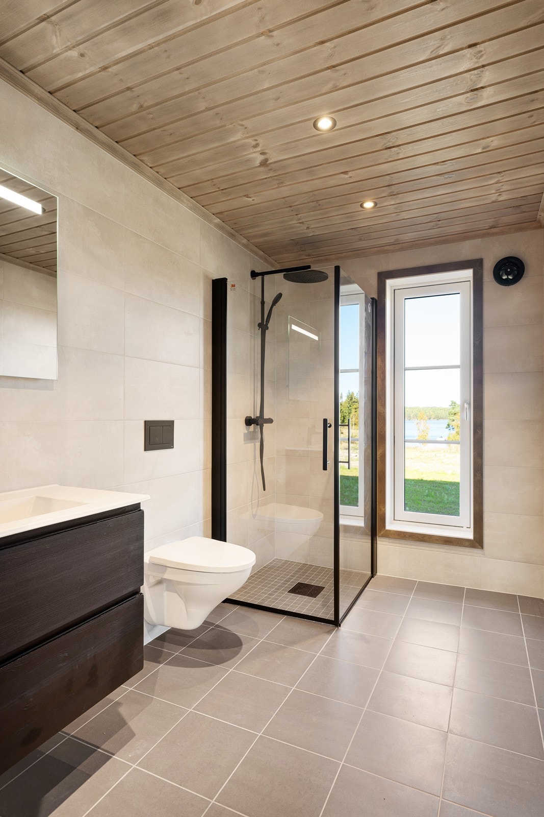 Badet er innredet med vegghengt toalett, dusjhjørne med innfellbare glassvegger, servant i baderomsinnredning og speil med belysning. Galleribilde