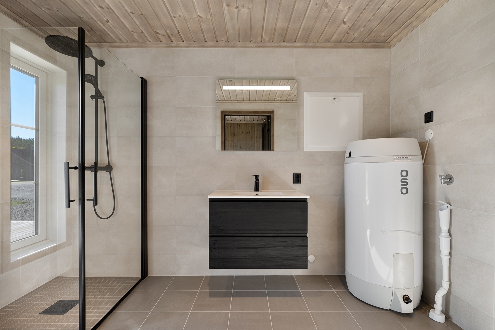 Bad/vaskerom med flislagte overflater. Badet er innredet med dusjhjørne med regnfallsdusj, servant i baderomsinnredning, vegghengt toalett, speil med belysning og opplegg for vaskemaskin. Galleribilde