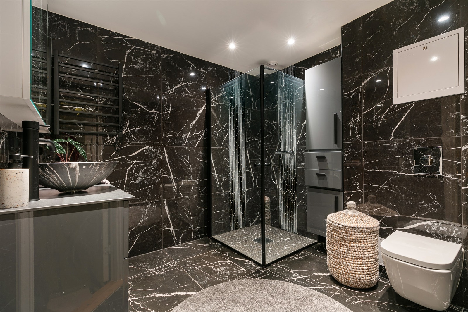 Moderne baderom med mørke fliser, hjørnedusj med glassvegger og vegghengt toalett. Stilrene detaljer og god oppbevaringsplass gir et eksklusivt preg. Galleribilde