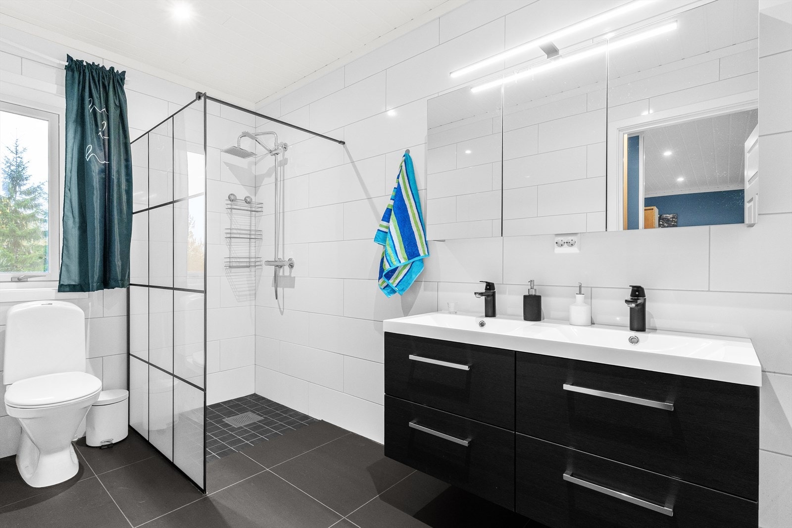 Lyst og pent innredet bad i tilknytning til hovedsoverommet med sort innredning med dobbelservant, oppbevaring i skuffeseksjon og speil med belysning. Videre er badet innredet med dusjhjørne med regnfalldusj og toalett. Galleribilde