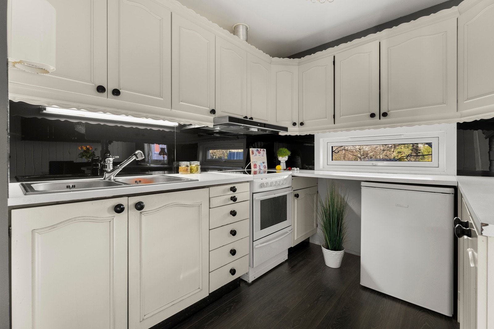 Eldre men velholdt kjøkkeninnredning med malte slette fronter og benkeplate av laminat. Kjøkkenet har nedsenket vask og nisjer med opplegg og avsatt plass til komfyr og kjøleskap. Galleribilde