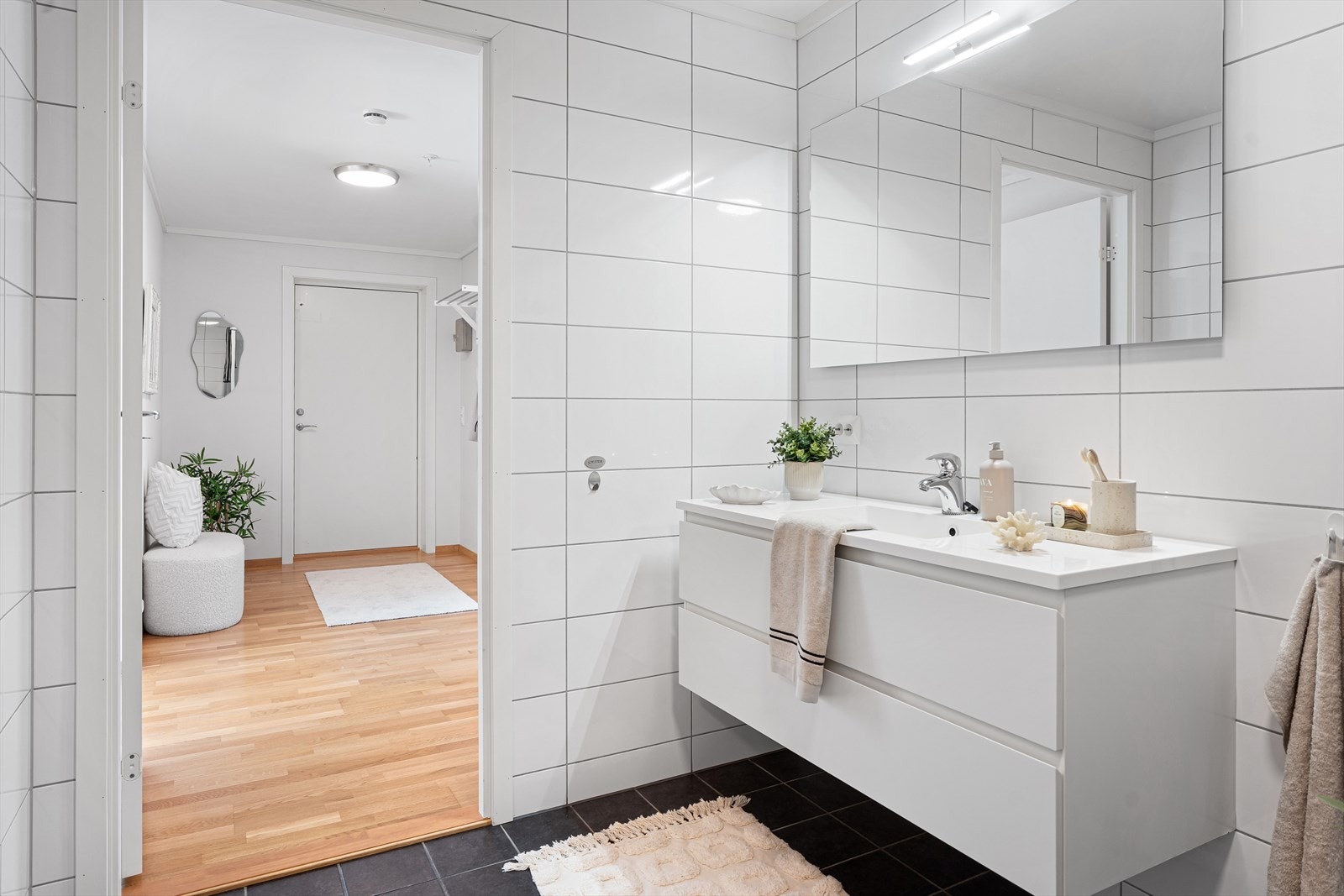 Badet er innredet med pen baderomsinnredning med hvite fronter, stort speil med belysning på vegg, vegghengt toalett og dusjkabinett Galleribilde